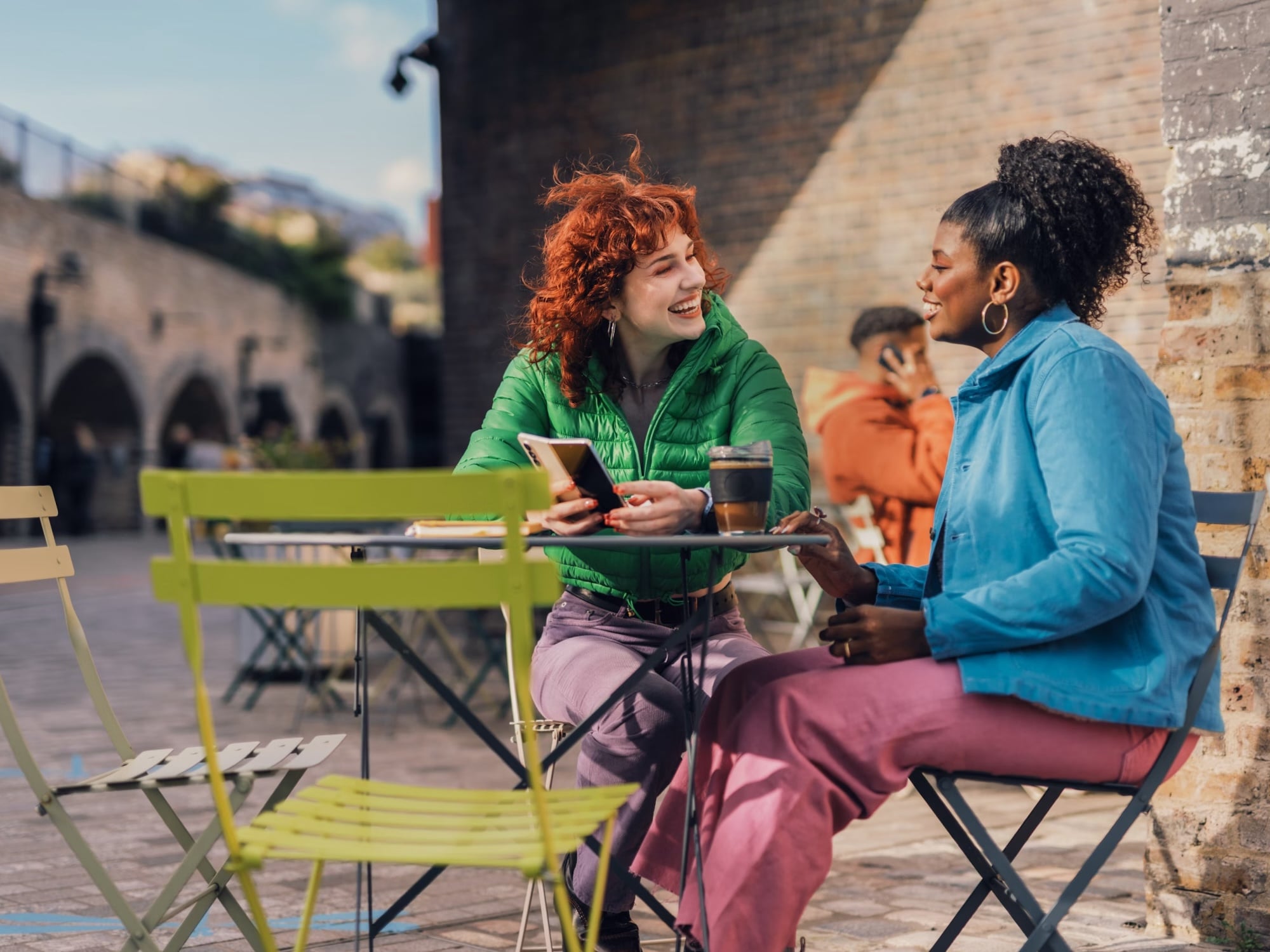 Two women chatting and laughing at an outdoor caf&eacute; table with drinks and bowls